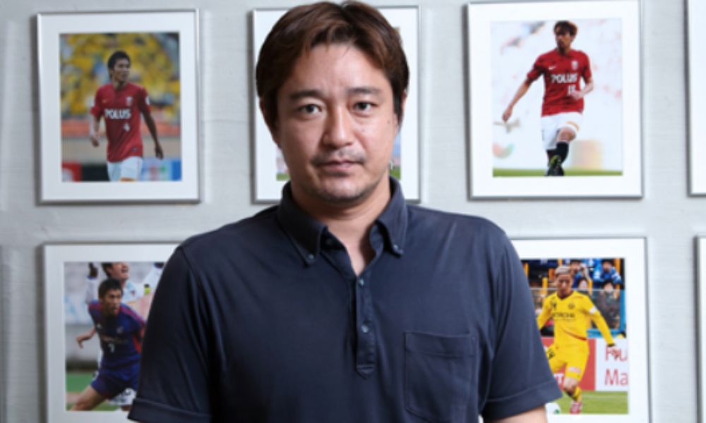 西塚定人氏(スポーツコンサルティングジャパン取締役)のポートレート。背景にサッカー選手の写真が飾られた壁前で撮影