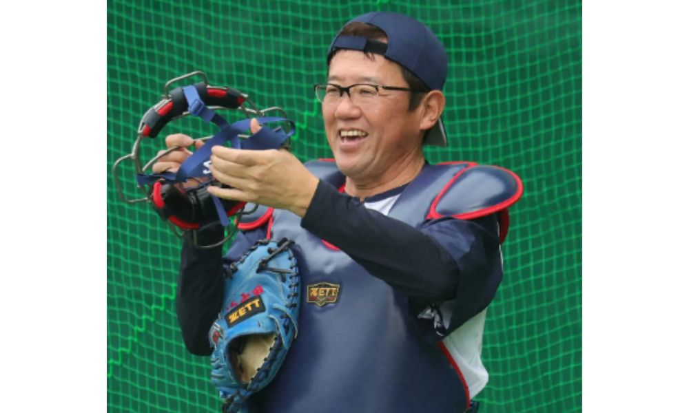 古田敦也さんがキャッチャーミットとマスクを手に笑顔でポーズをとる写真